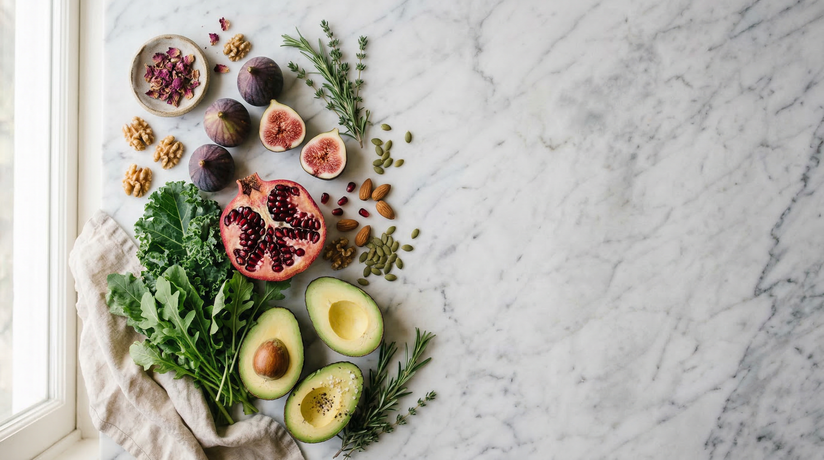 Artfully arranged fresh whole foods on marble — figs, pomegranate, avocado, herbs, and seeds